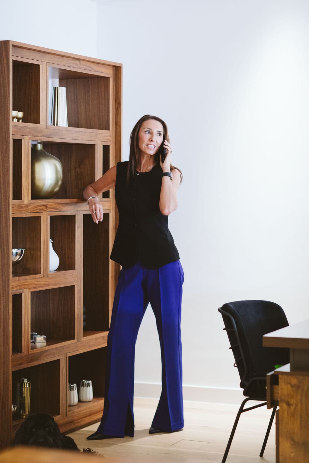 Woman in black vest and blue trousers talking on a phone by a wooden bookshelf.